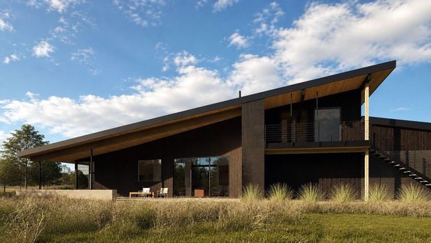 Exterior view of a modern two-level residence with a dramatic single-slope roof, dark vertical cladding, exposed timber soffits, extensive glazing, an elevated upper balcony, and an exterior stair. The house sits in a grassy rural-looking landscape beneath a partly cloudy sky.