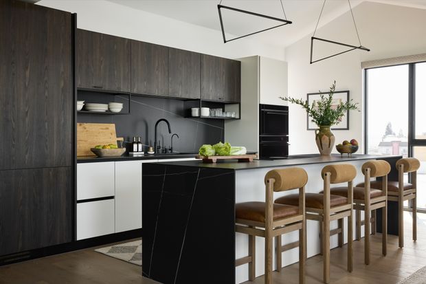A contemporary open-plan kitchen and dining area with dark wood full-height cabinetry, a matte black island and splashback, light benchtops, and a row of timber dining stools. Large glazing brings in natural light, while geometric black pendant lights and restrained styling create a refined, modern interior.