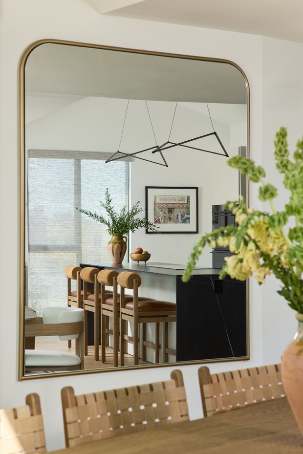 A large rounded-rectangle mirror with a thin brass-toned frame dominates the image, reflecting a refined open-plan dining and kitchen area. The reflection shows a long timber dining table with woven timber-back chairs, a dark kitchen island or cabinetry, upholstered counter stools, light-filtering window blinds, a framed artwork, greenery, and a sculptural black pendant light. The foreground also includes the dining table, chairs, and a large floral arrangement. The palette is warm and neutral with timber, cream, black, and brass accents.
