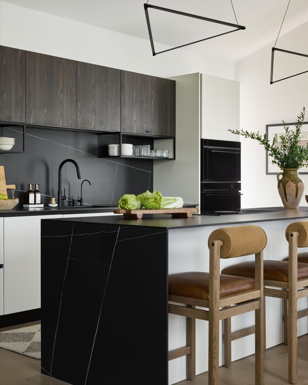 A refined contemporary kitchen with a large black stone island and integrated seating. Dark timber cabinetry, a charcoal splashback, pale lower cabinets, black appliances and minimalist suspended geometric pendant lights create a sophisticated monochromatic scheme, softened by timber stools, leather upholstery and greenery.