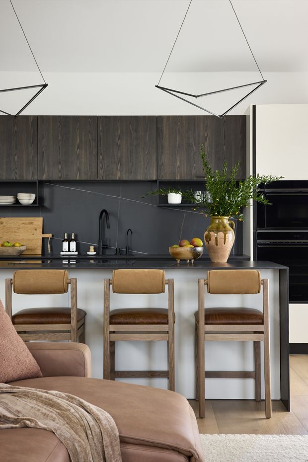 A refined open-plan kitchen and living area with dark vertical timber cabinetry, a charcoal stone island benchtop, integrated black appliances, and three timber-and-leather bar stools. Sculptural pendant lights hang above the island, while a neutral-toned sofa and rug occupy the foreground. Warm wood, leather, black finishes, and greenery create a sophisticated contemporary interior.