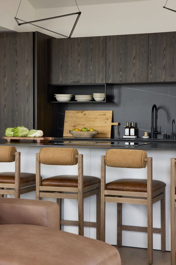 A refined contemporary kitchen with a dark wood-grain wall of overhead and full-height cabinetry, matte dark grey splashback, pale island cabinetry, and a dark benchtop. A raised breakfast bar is paired with timber-framed tan leather bar stools, while a large brown leather sofa occupies the foreground. Minimal black linear pendant lights, open shelving with white crockery, a timber chopping board, and a black gooseneck tap complete the warm, sophisticated interior.