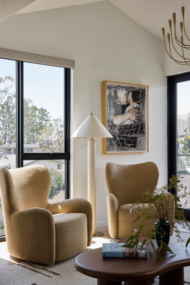 A refined living room flooded with natural light through large black-framed windows. Two sculptural boucle lounge chairs sit around a rounded dark timber coffee table, with a tall floor lamp, framed artwork, soft neutral walls, and a large textured rug. The room has a vaulted ceiling and an elegant brass multi-arm chandelier, creating a warm, contemporary and inviting atmosphere.