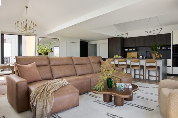 Open-plan contemporary living and kitchen area with a large tan leather sectional sofa and ottoman, sculptural timber coffee table, patterned area rug, and soft neutral furnishings. A dark timber kitchen with island seating, integrated appliances, pendant lights, and black-framed glazing occupies the background. The room is bright, refined, and connected to an outdoor balcony or terrace.