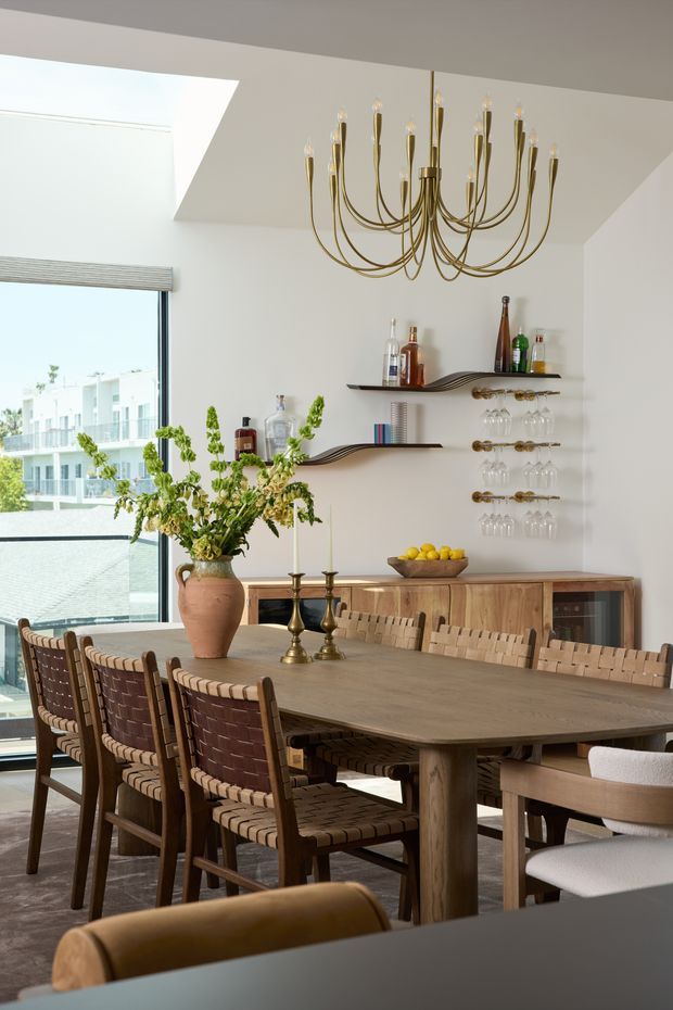 A refined dining room with a large natural timber dining table surrounded by woven leather and timber dining chairs. A sculptural brass chandelier hangs above, while a timber sideboard, decorative open shelving, hanging wine-glass racks, and bottles create a sophisticated bar display. Floor-to-ceiling glazing brings in abundant daylight and overlooks a modern urban setting. The palette is warm and neutral, with white walls, timber, brass, and earthy brown furnishings.