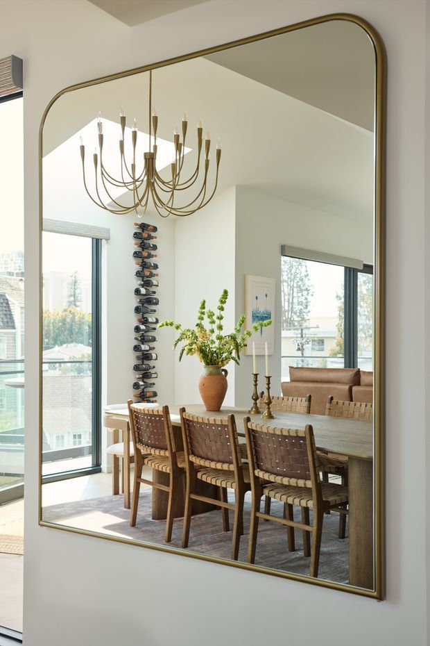 A large gold-framed wall mirror reflects a bright dining room with a long wooden dining table, woven leather dining chairs, a sculptural chandelier, a tall decorative wine-bottle display, large windows, and a nearby living area. The image is focused on the refined dining interior as seen through the mirror.