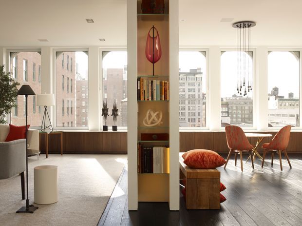 Bright, high-rise apartment interior with an open-plan living and dining area. Floor-to-ceiling windows provide expansive urban views, while a central built-in shelving partition separates the spaces. The living area includes a neutral sofa, floor lamps, a red accent cushion, a dark timber window seat and pale carpet. The dining area has a timber table, upholstered rust-orange chairs, dark timber flooring, and a sculptural pendant light. The space has clean contemporary lines and abundant natural light.