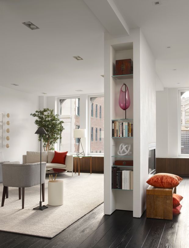 Bright, refined contemporary living room in an urban apartment, featuring white walls and ceiling, expansive windows, dark timber flooring, a pale area rug, upholstered lounge chairs, a sofa with red cushions, and a built-in open shelving partition displaying books and decorative objects. A compact seating nook with a timber cabinet and orange-red cushions is visible beside the partition.