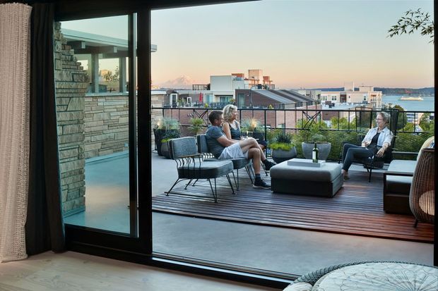 Interior view looking through large black-framed sliding glass doors onto a furnished rooftop terrace. The terrace has timber decking, outdoor lounge seating, potted plants, and a small table, with urban waterfront buildings and distant mountains beyond. The interior foreground includes pale timber flooring, curtains, and a partial upholstered seat.