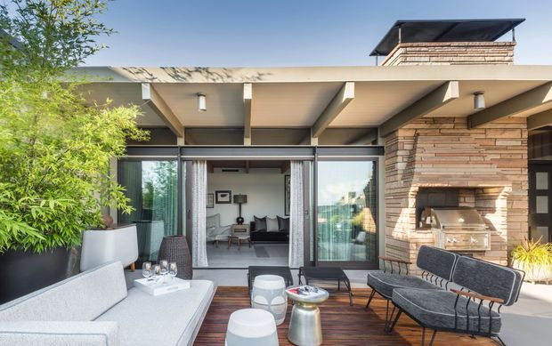 A contemporary outdoor terrace adjoining a glazed living room. The space features a dark timber deck, built-in pale upholstered seating, a freestanding lounge chair and stools, a timber-clad or rendered ceiling with exposed beams, and a substantial horizontal brick feature wall incorporating a built-in barbecue. Sliding glass doors connect the terrace to the interior living area, with lush potted planting at the edges.