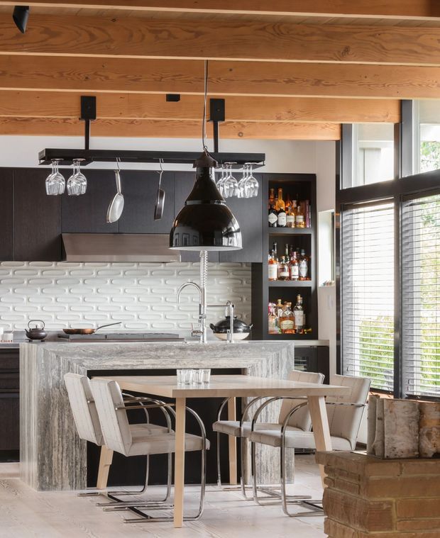 A sophisticated open-plan kitchen and dining area with a large stone island, integrated sink and tapware, dark cabinetry, a white textured tile splashback, and a built-in open bar display. A timber-lined ceiling adds warmth, while a black linear pendant with hanging glassware and a sculptural pendant light provide strong focal points. The island extends into a dining table surrounded by upholstered cantilever chairs. Large windows with dark frames and blinds bring in abundant natural light.