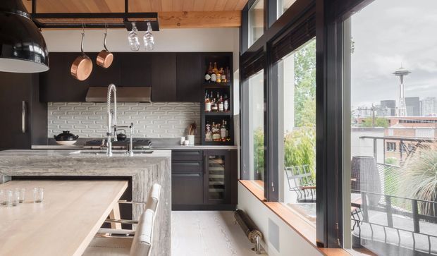 A contemporary kitchen with dark built-in cabinetry, a large grey stone island, white textured brick-style splashback, and exposed timber ceiling beams. Pendant lights and copper cookware hang above the island. Full-height black-framed glazing opens the room visually to an outdoor balcony and an urban skyline, while a timber-lined ceiling and warm wood window sill add warmth.