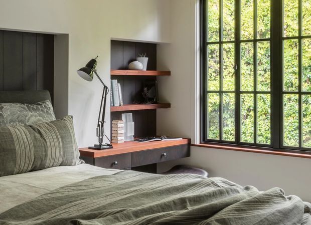 A calm, contemporary bedroom with a dark built-in bedhead, integrated open shelving and a compact desk in warm reddish timber, dark cabinetry, and a black articulated task lamp. Layered muted green bedding and cushions create a soft, coordinated palette. A large black-framed multi-pane window brings in abundant natural light and views of greenery.