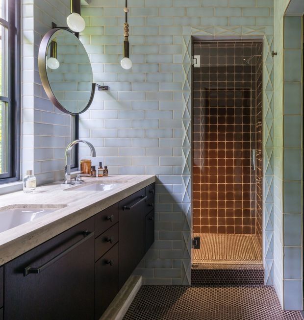 A refined bathroom with a long dark built-in vanity, pale marble benchtop, undermount basin, wall-mounted oval mirror, and contemporary pendant lights. The walls are clad in pale blue-green rectangular tiles with geometric textured tile detailing around the shower opening. A walk-in shower features warm brown square wall tiles and a small hexagonal mosaic floor, continuing across the bathroom floor. A large window provides natural light.