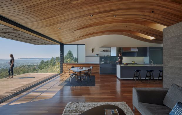 Open-plan contemporary living, dining and kitchen space with expansive glass sliding doors opening onto a timber terrace and panoramic landscape views. The interior features a dramatic curved timber-lined ceiling, polished dark timber flooring, dark built-in kitchen cabinetry, a light island bench, and an exposed textured masonry wall. A dining table and lounge seating occupy the foreground, while strong sunlight casts linear shadows across the floor and deck.