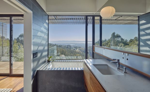 A contemporary bathroom with a freestanding rectangular bathtub positioned beside floor-to-ceiling glazing, overlooking a wooded landscape and distant water. The room features a long grey stone vanity with integrated basins, warm timber cabinetry, dark blue-grey wall tiles, slim dark window frames, and a large spherical pendant light. Exterior sun-shading creates strong striped shadows across the floor, tub, and walls.