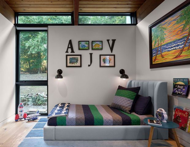 A compact, brightly lit bedroom with a built-in upholstered bed beneath a high clerestory window. The room features white walls, a warm timber-lined ceiling with exposed beams, tall black-framed glazing overlooking greenery, colourful framed artwork and letter decorations, and a striped quilt in green, purple, blue and neutral tones. A small round side table and children's books and toys suggest a playful bedroom setting.