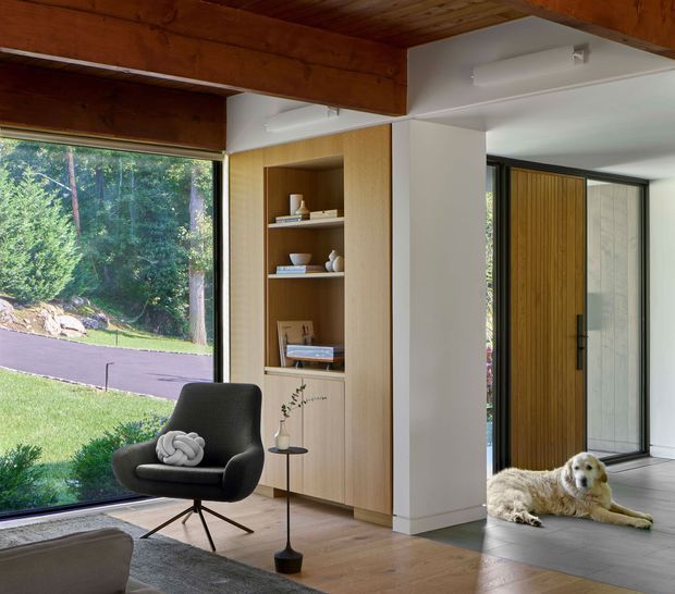A warm, modern living room with expansive glazing overlooking a landscaped yard, a dark lounge chair, built-in light timber shelving, and timber ceiling beams. The space opens toward a tiled entry or circulation area with a tall timber pivot door, creating a strong indoor–outdoor connection. Natural daylight, restrained furnishings, and wood detailing give the room a calm contemporary character.