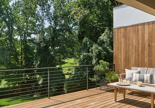 A sunny timber deck or terrace overlooking a lush, mature garden. The space features horizontal metal cable railing, warm timber decking, a wood-clad exterior wall with a white upper section, and a relaxed outdoor seating area with a sofa, cushions, coffee table, and potted plants.