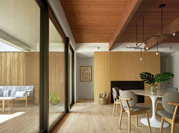 Warm contemporary open-plan dining area with a timber-lined ceiling, exposed timber beam, light timber flooring, and extensive dark-framed glazing along one side. A built-in light timber joinery wall incorporates a dark horizontal feature, while a round dining table and upholstered timber dining chairs sit beneath a minimalist suspended pendant light. A small adjacent lounge or sunroom is visible through the glazing.
