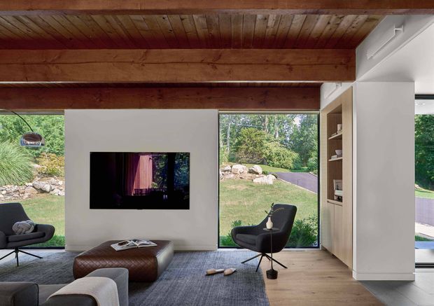 A refined contemporary living room with a warm timber ceiling and exposed structural beams, white plaster walls, large windows overlooking landscaped grounds, and built-in shelving. The room contains mid-century-inspired lounge chairs, a low upholstered ottoman, a television, and a large grey rug. Natural light and timber finishes create a warm, calm atmosphere.