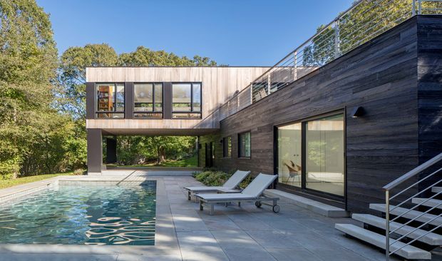 Contemporary multi-level residence with dark timber-clad walls, expansive glazing, and a cantilevered upper section surrounding a paved pool terrace. A rectangular swimming pool, loungers, stainless-steel balustrades, and exterior stairs define the outdoor setting, with mature trees beyond the property.