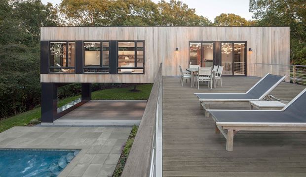 Exterior view of a modern elevated timber-clad house surrounded by trees. The building has a long rectangular form raised over a paved area, dark-framed windows, an outdoor dining area, and a broad timber deck with sun loungers overlooking a swimming pool.