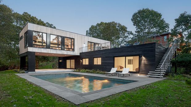 Contemporary multi-level residential home set in a landscaped, wooded site, photographed from the pool terrace. The house combines a dark horizontal-clad lower volume with a lighter upper box that projects outward, extensive glazing, rooftop terrace railings, and an external stair. A rectangular swimming pool with paved surround occupies the foreground, while warm interior lighting creates an inviting evening atmosphere.
