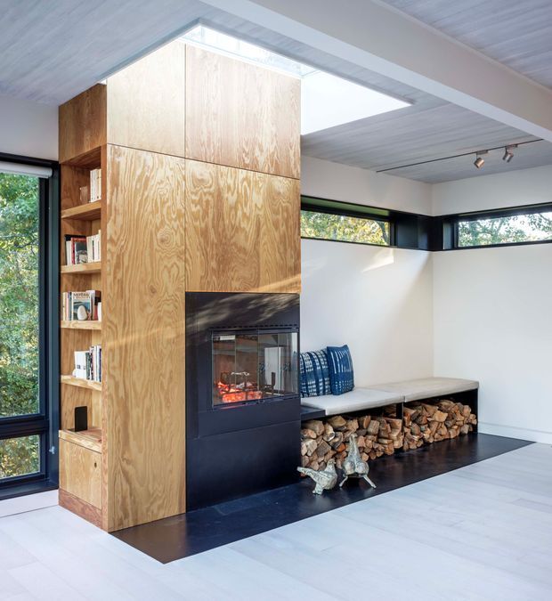 Bright contemporary living room centred on a tall plywood-lined fireplace and integrated open bookshelf. The black fireplace surround contrasts with a built-in window seat and exposed firewood storage, while clerestory glazing, corner windows and a skylight bring in abundant natural light. The room has pale timber flooring and a white painted ceiling with recessed timber lining and track lighting.
