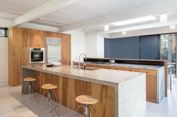 Bright contemporary kitchen with a large light stone island and integrated sink, timber veneer cabinetry, stainless-steel appliances, and a dark blue built-in cabinetry wall. The room features a pale timber-lined ceiling, exposed white beams, skylights, pendant lighting, and bar seating along the island.