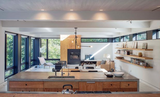 Bright open-plan kitchen and living area viewed across a large timber kitchen island. The room features extensive dark-framed glazing, white walls and ceiling, timber cabinetry and shelving, a fireplace set into a warm timber feature wall, and a lounge area beyond. The interior has a clean contemporary character with abundant natural light and integrated built-in joinery.