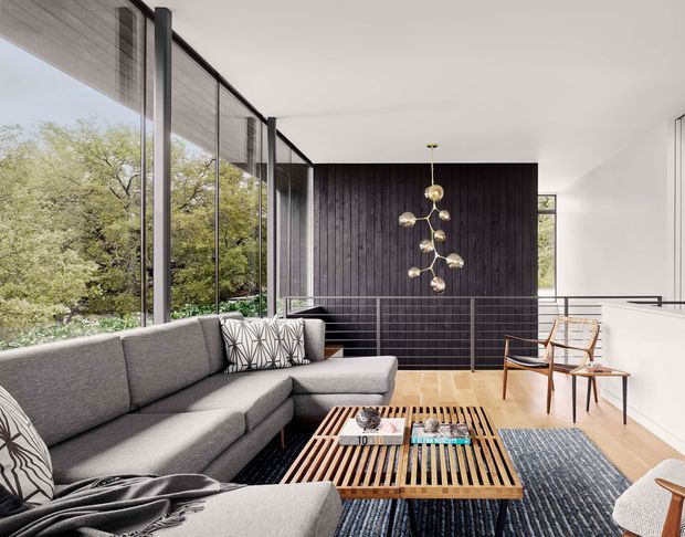 A bright contemporary living room with floor-to-ceiling glazing overlooking leafy trees. The space features a large grey sectional sofa, a slatted timber coffee table, pale timber flooring, a dark vertical timber feature wall, sculptural pendant lighting, and mid-century-style lounge chairs. A white stair or balustrade zone is visible beyond the seating area.