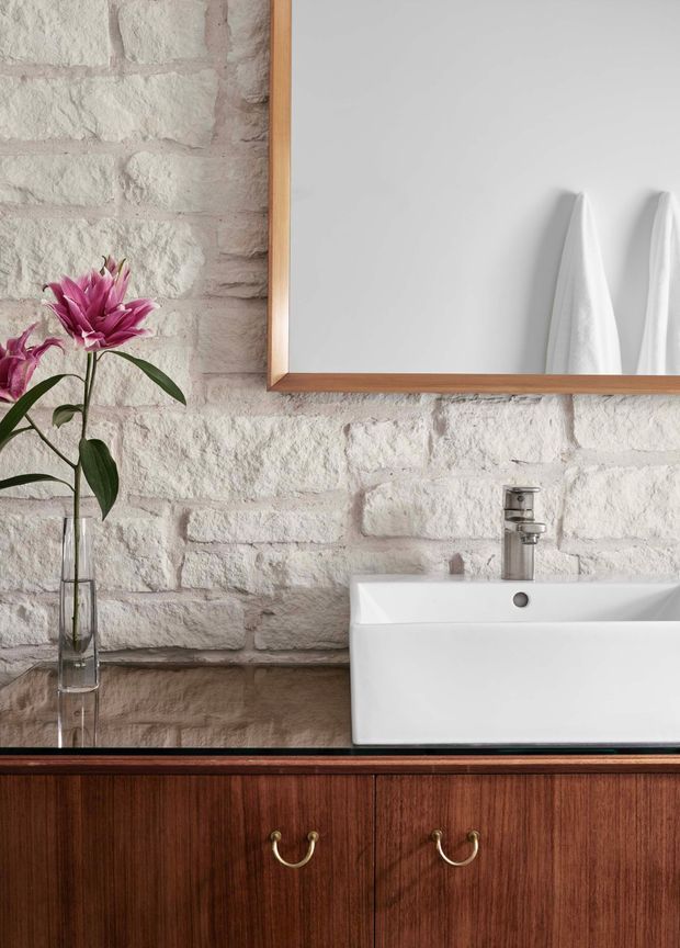 Close-up of a refined bathroom vanity featuring a white rectangular vessel basin with chrome tapware, dark timber cabinetry with brass pulls, and a white-painted exposed brick wall. A timber-framed mirror reflects neatly hung white towels, while a glass vase with a pink flower adds a small decorative accent.