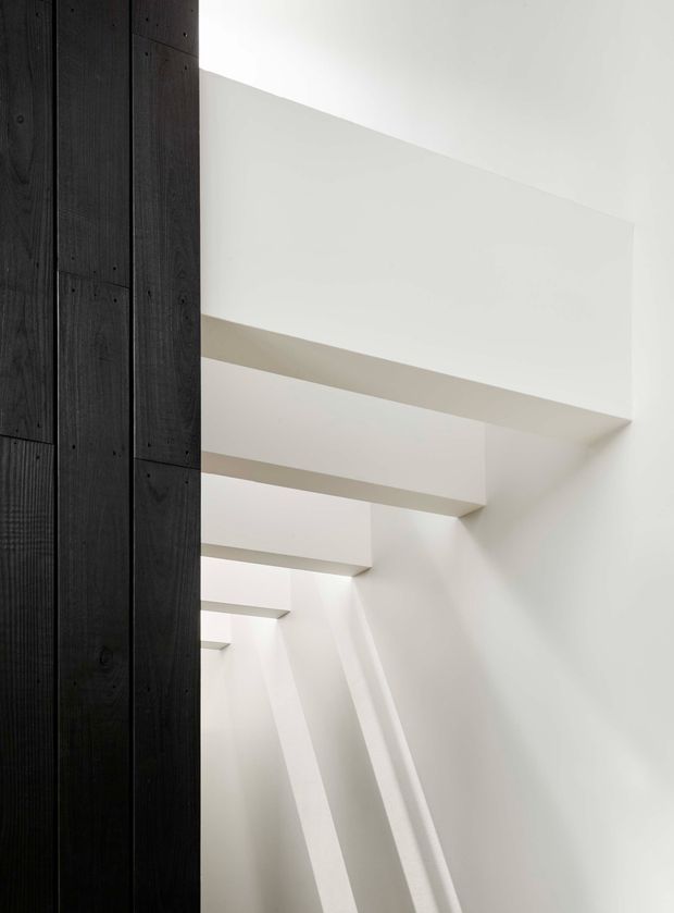 Minimal architectural interior detail showing a tall white stairwell or circulation void with repeated projecting horizontal landings or beams and strong angled daylight shadows. A contrasting vertical wall or screen of dark stained timber boards occupies the left side. The composition is geometric, sparse, and focused on light, shadow, and built form; no furnishings are visible.