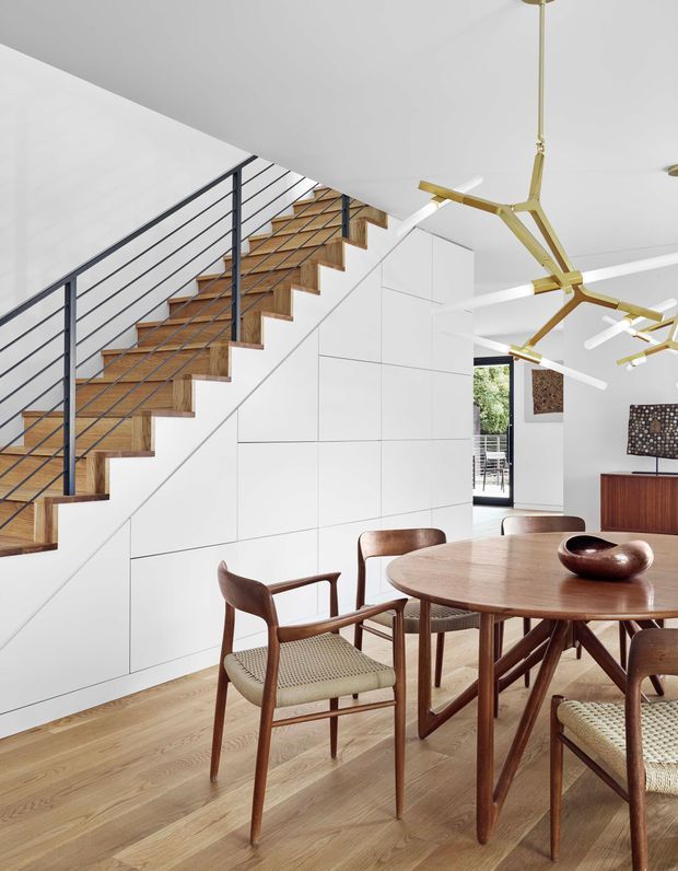 Bright open-plan dining area with a mid-century-inspired timber dining table and woven-seat chairs, adjacent to a sculptural white staircase. The staircase has warm timber treads, slim black metal balustrades, and white geometric wall cladding beneath it. White walls and ceilings, pale timber flooring, contemporary cabinetry, and branching brass pendant lights create a refined modern interior.