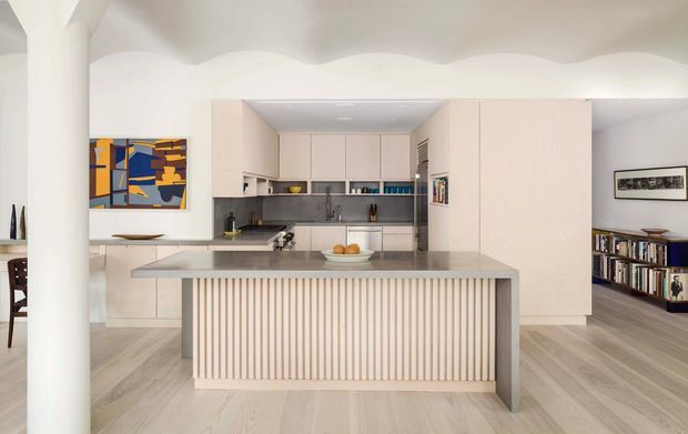 Bright open-plan kitchen dominated by a large island with a vertically fluted front, pale cabinetry, grey benchtops, and light timber flooring. A sculptural undulating ceiling, rounded structural column, open shelving, dining area, and adjacent living/storage zone are visible. The space has a refined contemporary, minimalist aesthetic with warm neutral finishes and colourful artwork as a small accent.