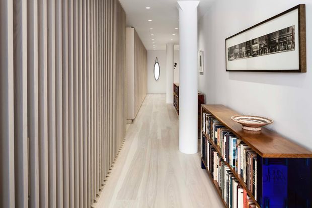 A long, bright contemporary hallway with pale timber flooring, a full-height vertical timber slat feature wall on the left, and a white cylindrical structural column near the centre. A low timber bookcase with books and decorative bowls lines the right wall beneath framed artwork. Recessed ceiling lights and a rounded wall mirror extend the view down the corridor.
