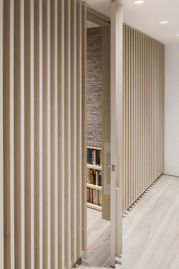 A minimalist interior corridor lined with closely spaced vertical light timber battens. The slatted screen forms a partially concealed doorway and reveals built-in bookshelves and a textured stone or brick feature wall behind it. Pale timber flooring, white walls, recessed ceiling lights, and warm neutral tones create a clean, refined architectural interior.