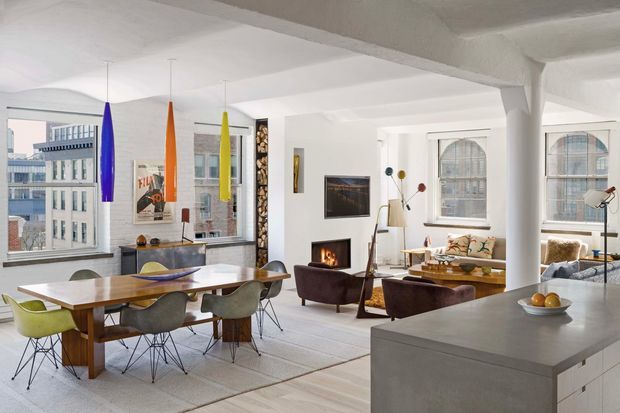 Bright open-plan loft interior with a dining area, living room and kitchen in a large industrial-style space. The room features expansive steel-framed windows, white-painted structural columns and beams, a suspended fireplace, timber dining table, modern dining chairs, and colourful pendant lights. A grey kitchen island occupies the foreground, while the living area includes a sofa, armchairs and a television.
