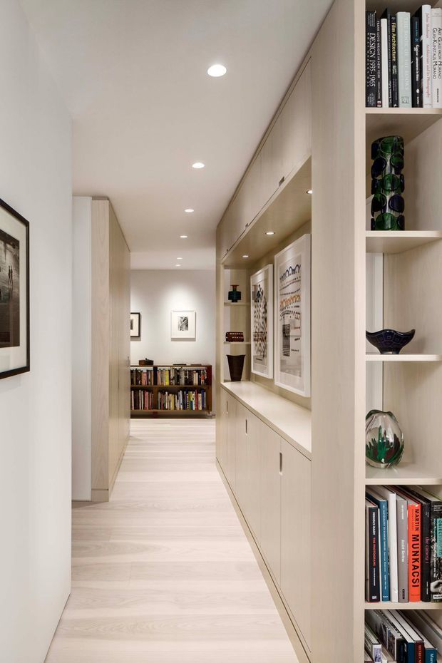 A narrow, well-lit residential hallway lined with pale timber built-in cabinetry and open bookshelves. The corridor features integrated display niches, concealed storage, framed artwork, recessed ceiling lights, and a low bookcase at the far end. Pale timber flooring and joinery create a calm, minimalist library-like passage.