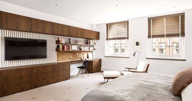 A bright, refined bedroom with a built-in walnut entertainment and study wall. The joinery includes vertical slatted detailing, open shelving, a desk, drawers, and a wall-mounted television. A bed, lounge chair, ottoman, and two windows with timber blinds occupy the room. White-painted brick walls and a pale patterned carpet create a warm contemporary interior.