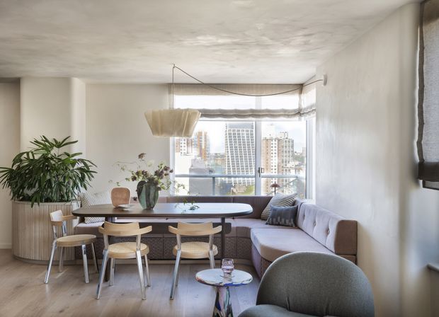 Bright, warm-toned open-plan dining and living area in an apartment, with a built-in window-side banquette, oval dining table, upholstered dining chairs, curved sectional sofa, sculptural pendant light, indoor plants, and broad city views through the glazing. The room has white textured walls and ceiling, light timber flooring, and a relaxed contemporary feel.