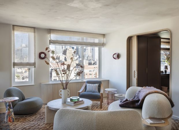 A bright, contemporary living room in an urban apartment with large windows overlooking a dense city skyline. The room features a textured plaster ceiling, pale walls, rounded boucle seating, a low round coffee table, patterned rug, sculptural side tables, and a dark timber-framed opening leading to another space. Roman blinds soften the daylight, while a radiator sits beneath the windows.