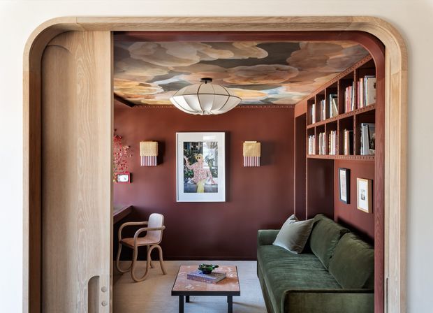 A richly decorated compact sitting room or reading lounge viewed through a wide rounded timber-framed opening. The room has deep reddish-brown walls, a dramatic painted cloud ceiling, built-in dark timber shelving filled with books, a green upholstered sofa, a small timber coffee table, a bentwood chair, framed artwork, wall lights, and a sculptural pendant. The composition feels intimate, theatrical, and highly crafted.