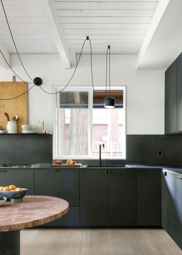 A contemporary kitchen with dark matte cabinetry arranged along two walls, a dark benchtop, integrated sink and cooktop, and a rounded peninsula or island with a pink-toned stone surface. The space features white painted timber plank ceilings with exposed beams, white walls, a timber backsplash panel, a large window, and black pendant lights with exposed cords. Light timber flooring and strong natural light create contrast with the charcoal joinery.