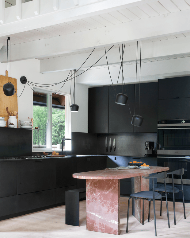 A contemporary kitchen with extensive matte-black cabinetry and appliances, a dark backsplash, pale timber flooring, and a sculptural pink stone island with an integrated pedestal base. Black pendant lights hang from long exposed cords beneath a white panelled ceiling. A window brings in greenery and daylight, while dark stools surround the island.