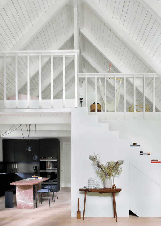 Bright double-height open-plan interior with a dramatic white pitched timber roof, exposed structural framing and mezzanine-level balustrades. A sculptural white staircase descends beside a compact black kitchen with integrated appliances, pendant lights and a pink stone island. Mid-century timber console styling and decorative objects sit beneath the stairs, while the upper level contains a hammock and other furnishings.