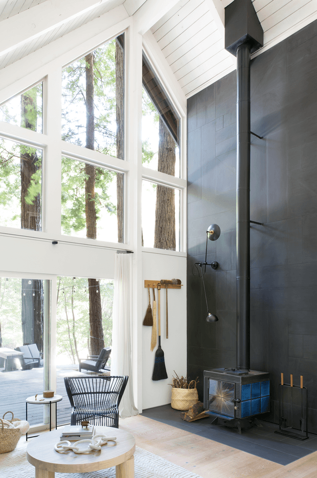 Bright, double-height living room in a woodland setting, featuring a dramatic wall of floor-to-ceiling glazing, white-painted timber ceiling boards and beams, and a dark tiled feature wall with a freestanding wood-burning stove and flue. The room has light timber flooring, a woven lounge chair, round coffee table, fireplace tools, and natural woven accessories. A deck and forest are visible through the glazing.