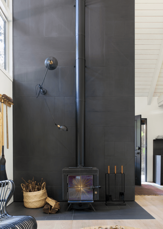 Interior view of a dramatic dark tiled feature wall with a freestanding wood-burning stove and tall black flue. A simple wall-mounted light, firewood basket, log holder and fireplace tools sit beside the stove. The room has a pale timber-lined ceiling, light timber flooring, and glimpses of an adjacent bright living area through an opening. The overall aesthetic is modern and industrial with strong dark-light contrast.