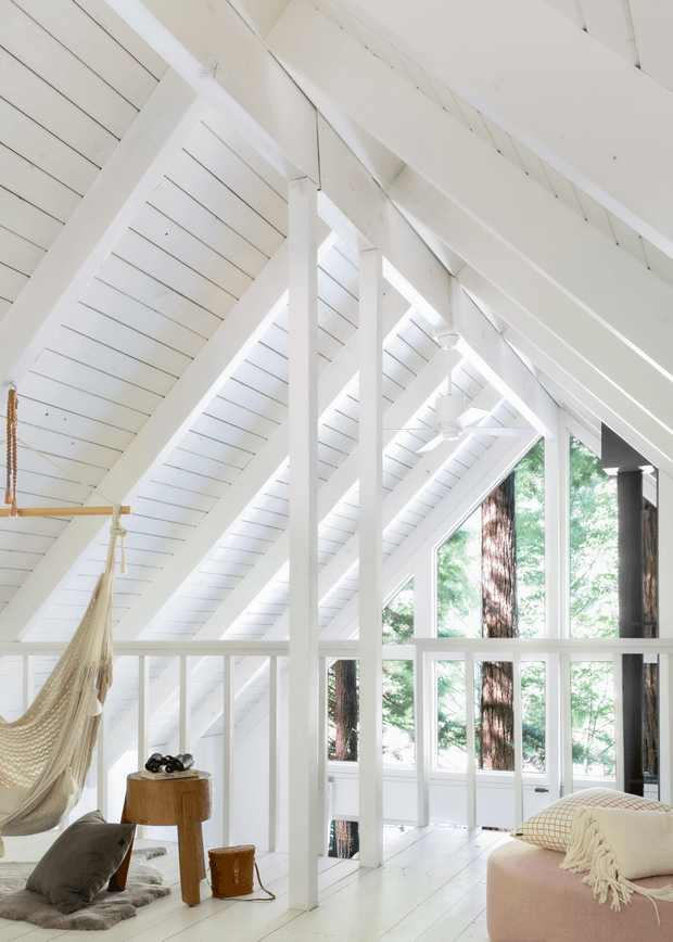 Bright, airy loft-like interior with dramatic white painted timber roof framing, exposed rafters, sloped ceiling planes, and large floor-to-ceiling windows overlooking trees. A hammock, low seating, side tables, cushions, and a soft upholstered bench create a relaxed retreat atmosphere. White timber flooring, balustrades, structural posts, and ceiling boards dominate the space, with ceiling fans and a dark vertical fireplace or column adding contrast.